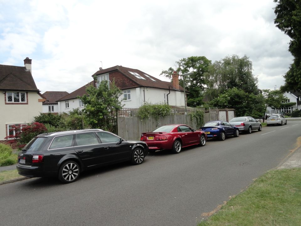 Several cars parked on a street in front of houses, including a black wagon, a red coupe, and a blue coupe