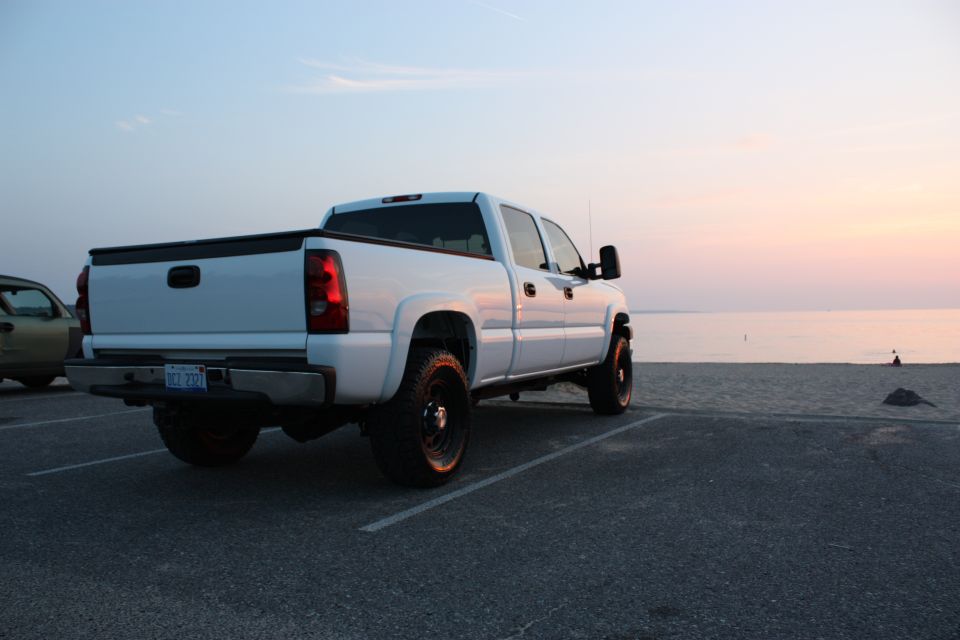 White pickup truck parked near beach at sunset. Another vehicle is partially visible to the left