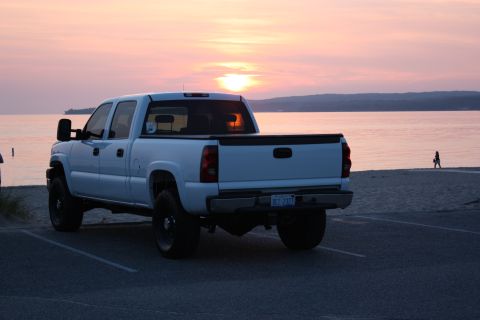 2006 Chevrolet Silverado 2500 - A white pickup truck is parked on asphalt near a beach as the sun sets over the water.