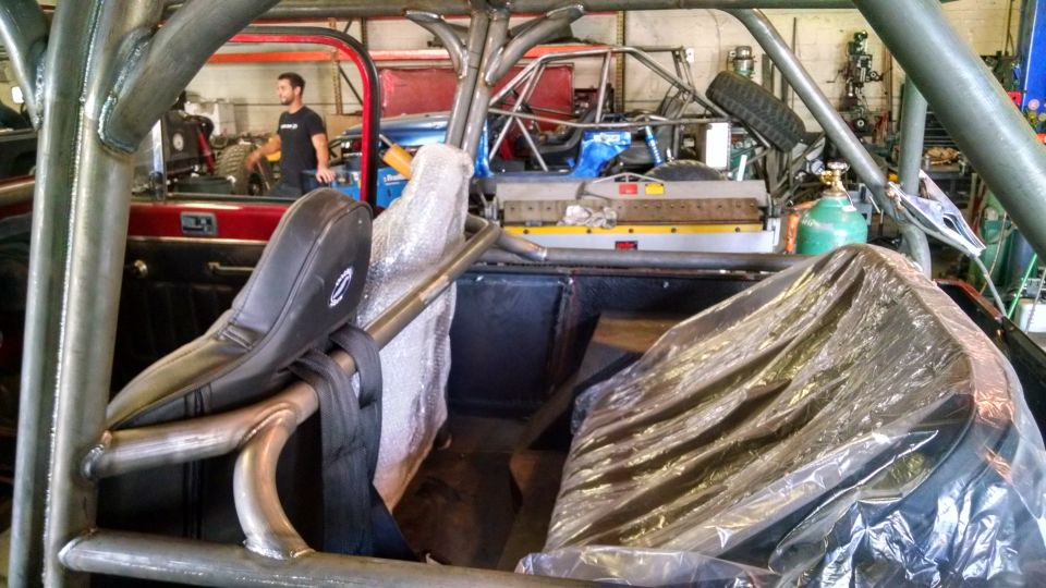 Interior view of a vehicle with a prominent roll cage, custom seats, and a person standing in the background near another vehicle