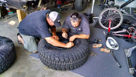 1985 Jeep CJ7 - Men assemble a tire and rim in a garage, with various tools and spare tires visible in the background.