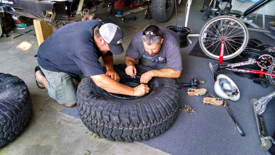Two men kneel on a garage floor, working on a large off-road tire and rim. Tools and other tires are scattered around