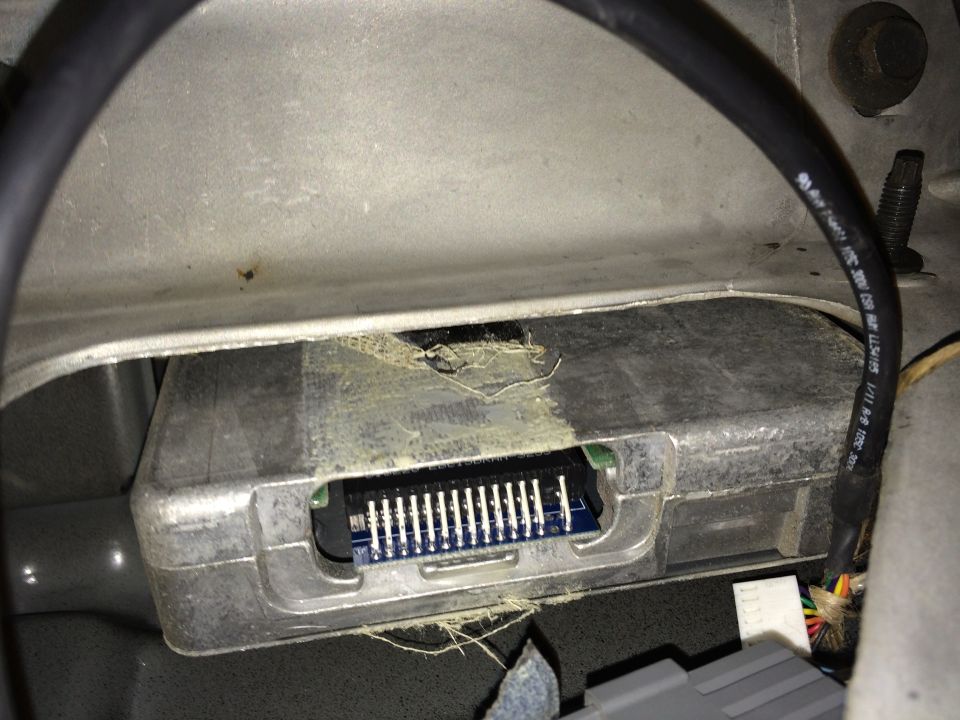 Close-up of an electronic control module with a multi-pin connector. Some yellow adhesive is visible around the module