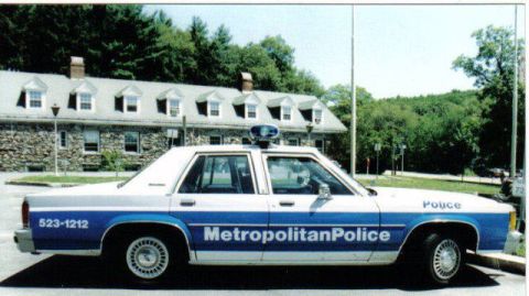 1991 Ford LTD Crown Victoria - A white and blue police car is parked in front of a stone building with trees in the background.