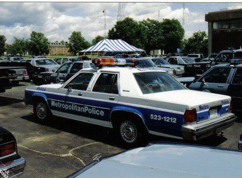 1991 Ford LTD Crown Victoria - A 1991 Ford Crown Victoria LTD police car is parked in a lot with other vehicles under a striped tent.