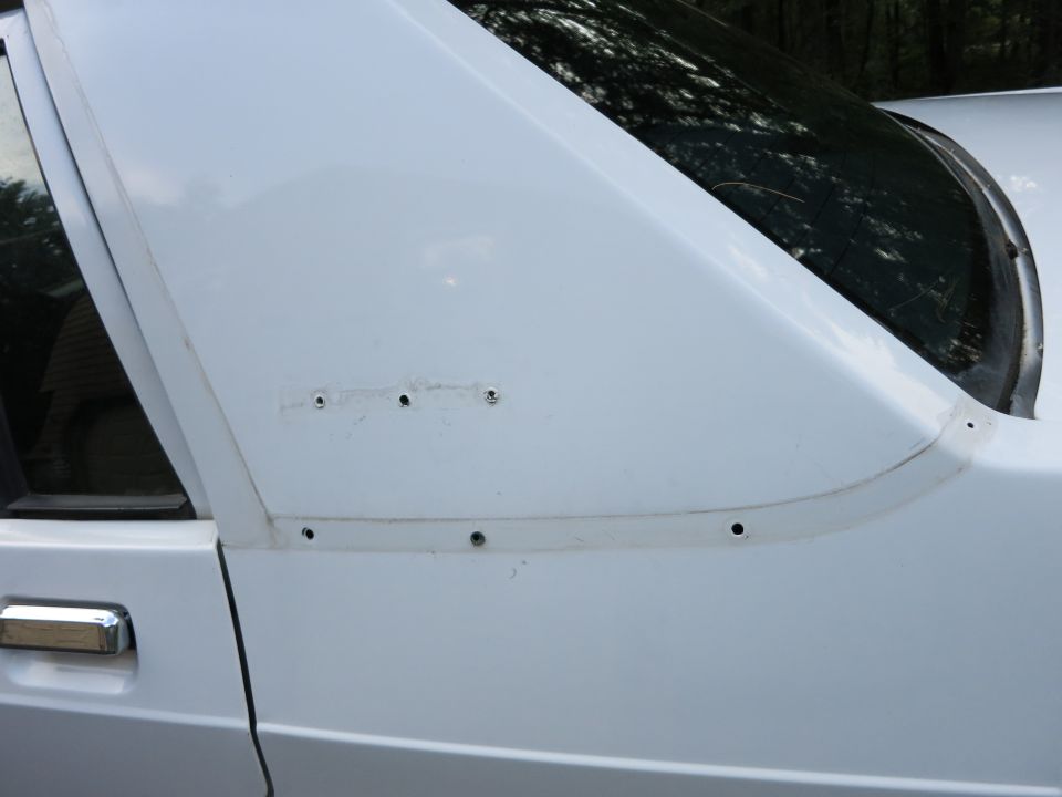 Close-up of a white car's rear door and C-pillar area, showing screw holes along a trim line
