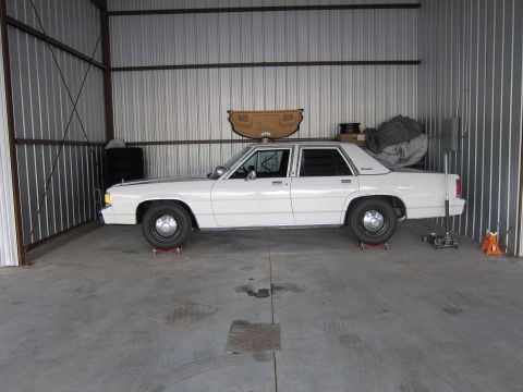1991 Ford LTD Crown Victoria - Side view of a white 1991 Ford Crown Victoria LTD sedan, possibly undergoing maintenance in a garage.
