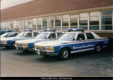 1991 Ford LTD Crown Victoria - A line of Metropolitan Police cars, likely from the early 1990s, are parked outside a building.