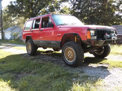 1986 Ford Bronco - This red Jeep Cherokee XJ is modified with an aftermarket bumper and winch, and sits on a gravel and grass area.