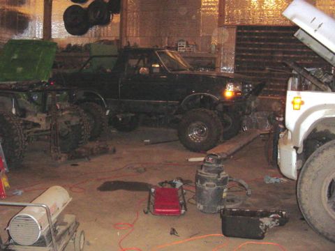 1992 Ford Bronco - Two trucks are in a garage, one is a dark green pickup and the other is a white truck chassis, with various tools and heaters around.