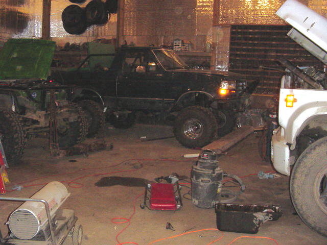 Dark green pickup truck with lifted suspension parked next to a white truck chassis in a garage. Tools and heaters are on the floor