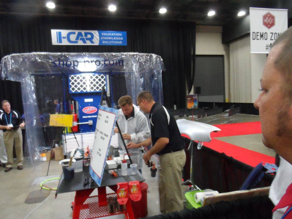 Automotive paint booth demonstration with two men working and a silver car fender on display