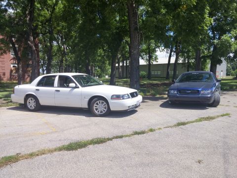 2000 Mercury Grand Marquis - Two sedans are parked outdoors on a sunny day, with trees and buildings in the background.