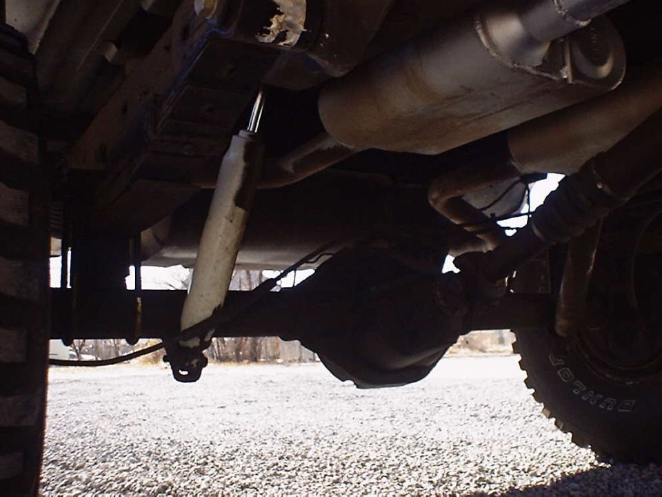 Underside view of a vehicle's rear axle, shock absorber, and exhaust system. Tire tread visible