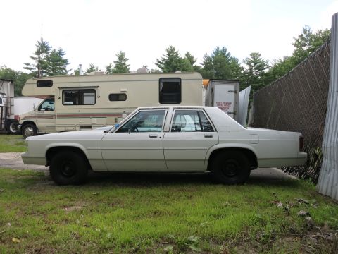 1991 Ford LTD Crown Victoria - A white 1980s Ford Crown Victoria is parked on grass next to an RV and a fence.