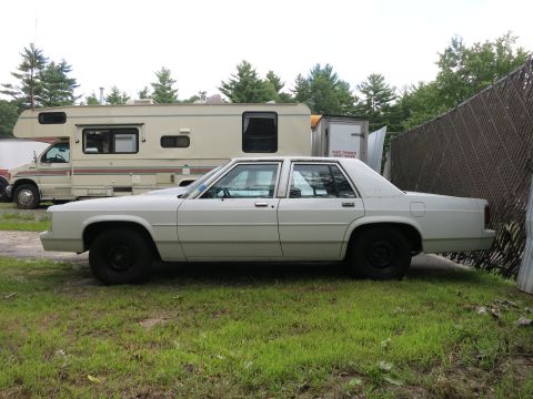 1991 Ford LTD Crown Victoria - A white sedan sits parked on grass next to a large RV, with a fence to its right.