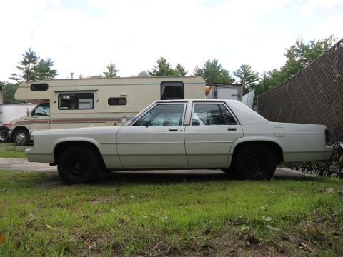 1991 Ford LTD Crown Victoria - White sedan parked on grass next to an RV, with a chain-link fence behind it.