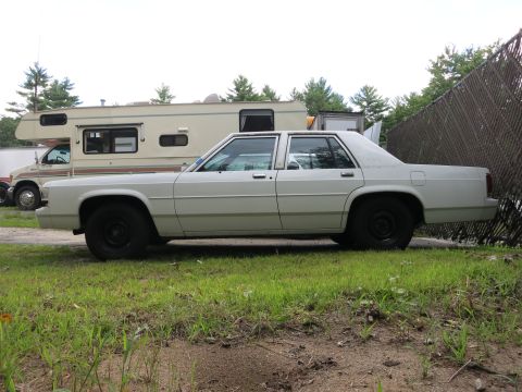 1991 Ford LTD Crown Victoria - A white sedan is parked in a grassy area next to an RV and a chain-link fence.