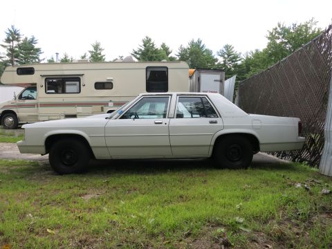 1991 Ford LTD Crown Victoria - A white sedan is parked on a grassy area next to a chain-link fence, with an RV visible behind it.