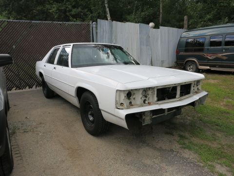 1991 Ford LTD Crown Victoria - A white sedan sits in a lot with its front bumper and grille missing, next to a parked van.