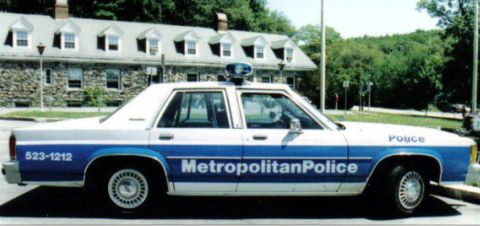 1991 Ford LTD Crown Victoria - A side view of a 1991 Ford Crown Victoria LTD police car parked in front of a stone building.
