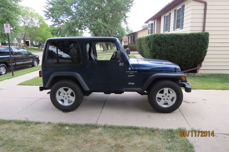 Side view of a dark blue Jeep Wrangler parked on a driveway. A black pickup truck is parked on the street