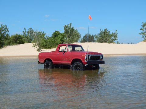 1978 Ford Bronco - A red pickup truck is partially submerged in water, with a sandy beach and trees behind it under a clear blue sky.