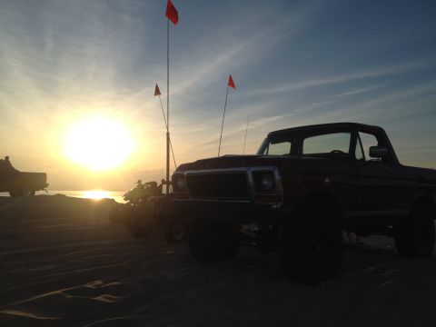 1978 Ford Bronco - A 1978 Ford Bronco sits on sand dunes as the sun sets over the ocean, with other vehicles and flags visible.
