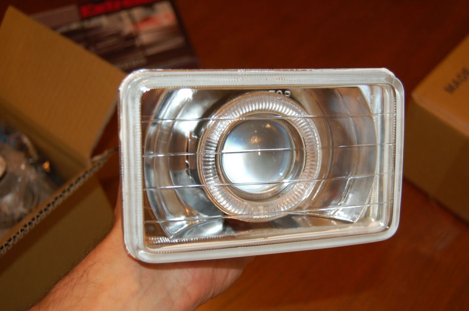 Hand holding a rectangular projector headlight with halo ring over cardboard boxes