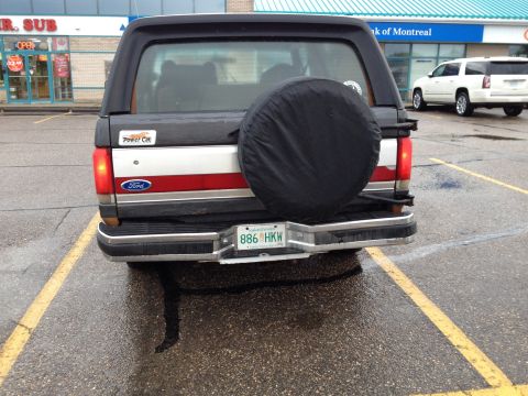 1989 Ford Bronco - A black Ford Bronco is parked in a lot, showing its rear end with a spare tire cover and a license plate.