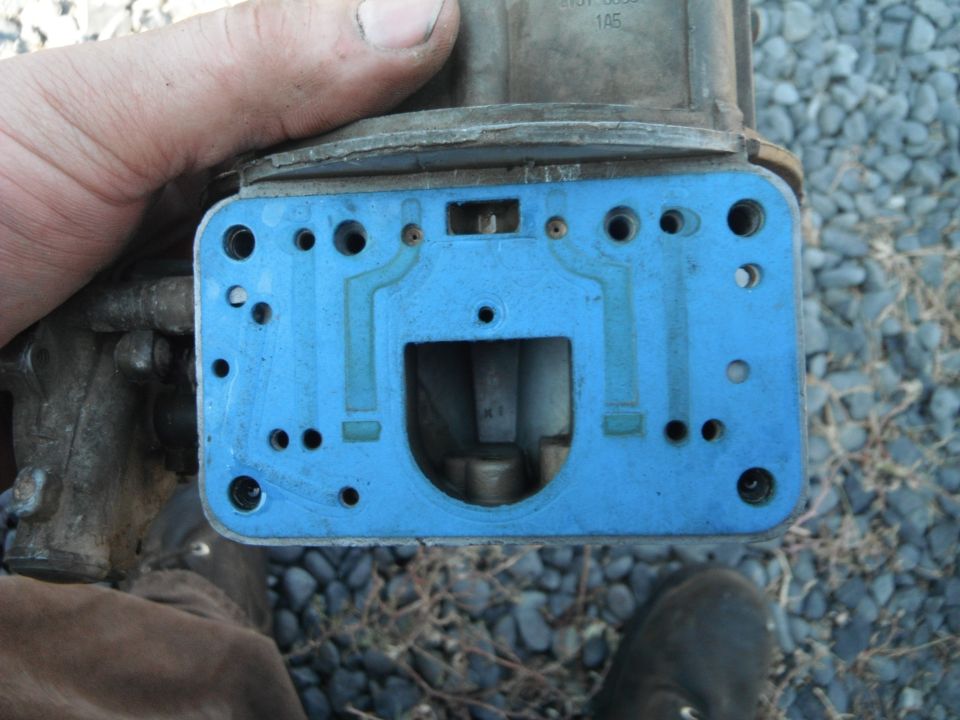 Close-up of a blue gasket on a carburetor base plate, held by a hand