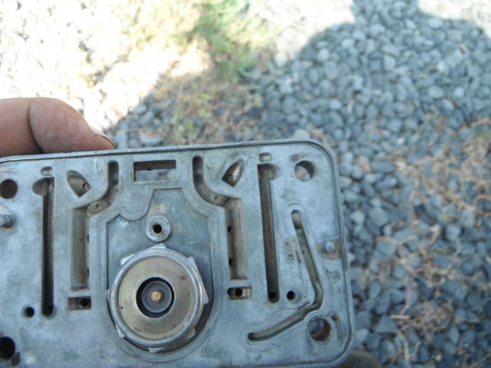 Close-up of a carburetor base plate with internal passages and a central brass fitting. A hand is visible at the top left