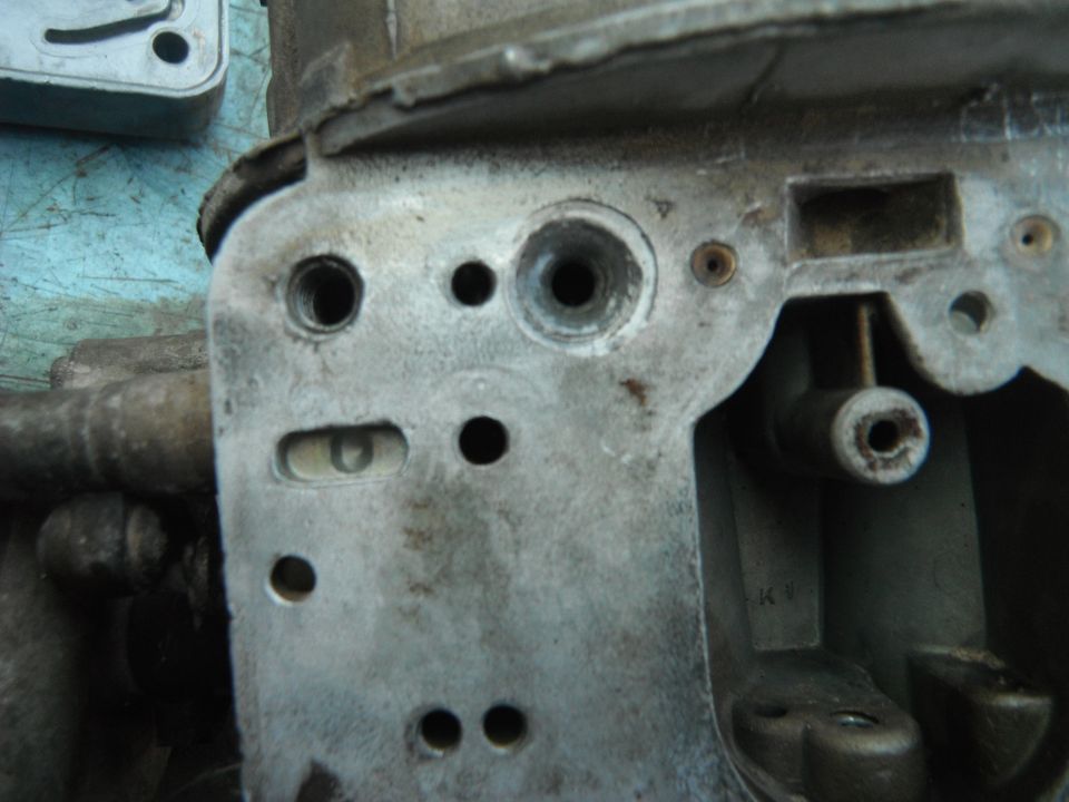 Close-up of a metal carburetor body with various holes and mounting points. Part of a Supermotors.net watermark is visible