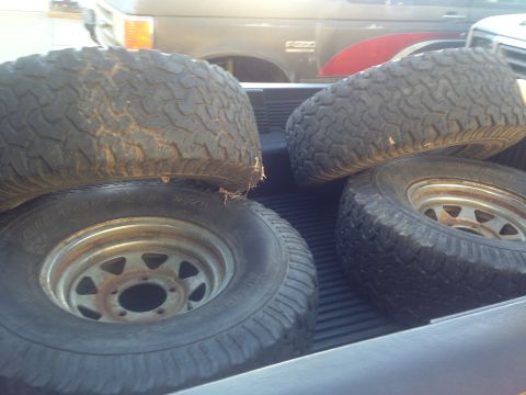 1986 Ford Bronco - Four off-road tires are loaded into the bed of a pickup truck, obscuring the view of the vehicle behind.