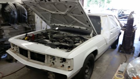 1991 Ford LTD Crown Victoria - A white sedan is undergoing repairs in a workshop, with its hood open to reveal the engine compartment.