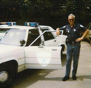 1991 Ford LTD Crown Victoria - Officer poses with a 1991 Ford Crown Victoria LTD police car, with another vehicle visible behind.