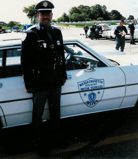 1991 Ford LTD Crown Victoria - Massachusetts Registry of Motor Vehicles police officer stands next to a marked patrol car, with other vehicles and officers in the backgro…