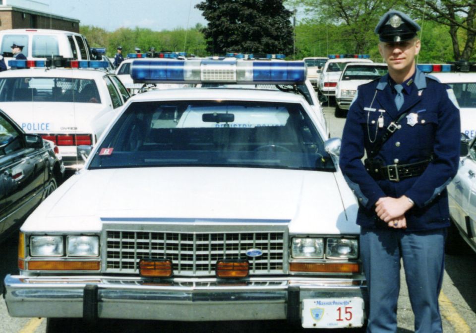 White 1991 Ford Crown Victoria Police Interceptor with blue light bar, front view. Officer stands beside it
