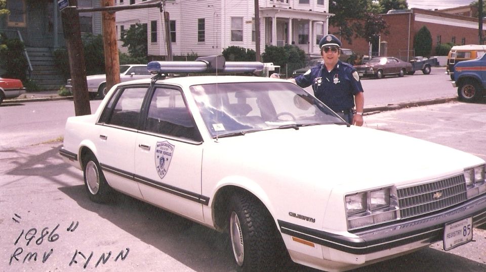 Officer standing next to a white 1986 Chevrolet Caprice police car. Text on car says 'Massachusetts Registry of Motor Vehicles'