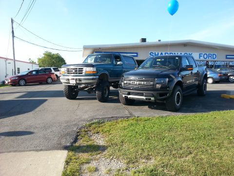 1996 Ford Bronco - A lifted 1996 Ford Bronco and a modern Ford F-150 Raptor are parked outside a Ford dealership.