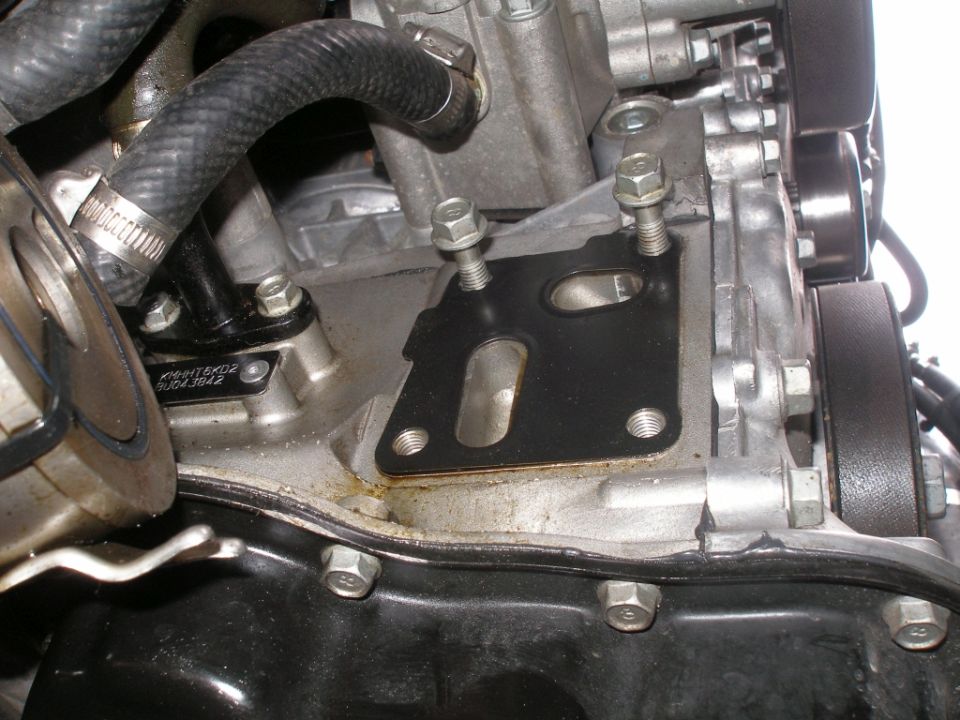 Engine block with gasket and bolts, showing a black gasket with two openings and metal bolts. A black hose is visible