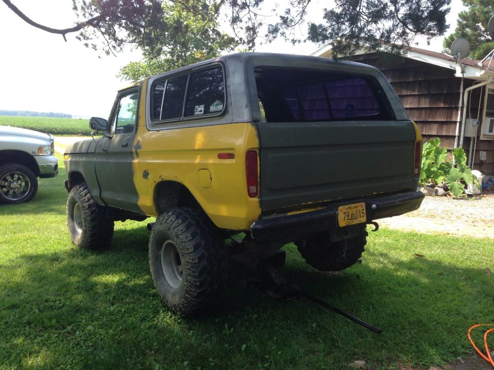 Rear view of a two-tone green and yellow Ford Bronco lifted on a jack. A silver pickup truck is parked to the left