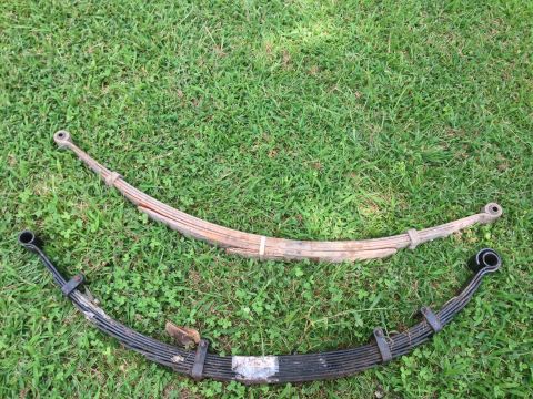 1979 Ford Bronco - A pair of leaf springs, one showing significant rust and the other painted black, are displayed on a grassy surface.