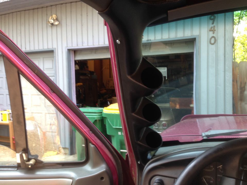 Maroon interior A-pillar with three black gauge pods. Visible through the windshield is a garage door with the number 940