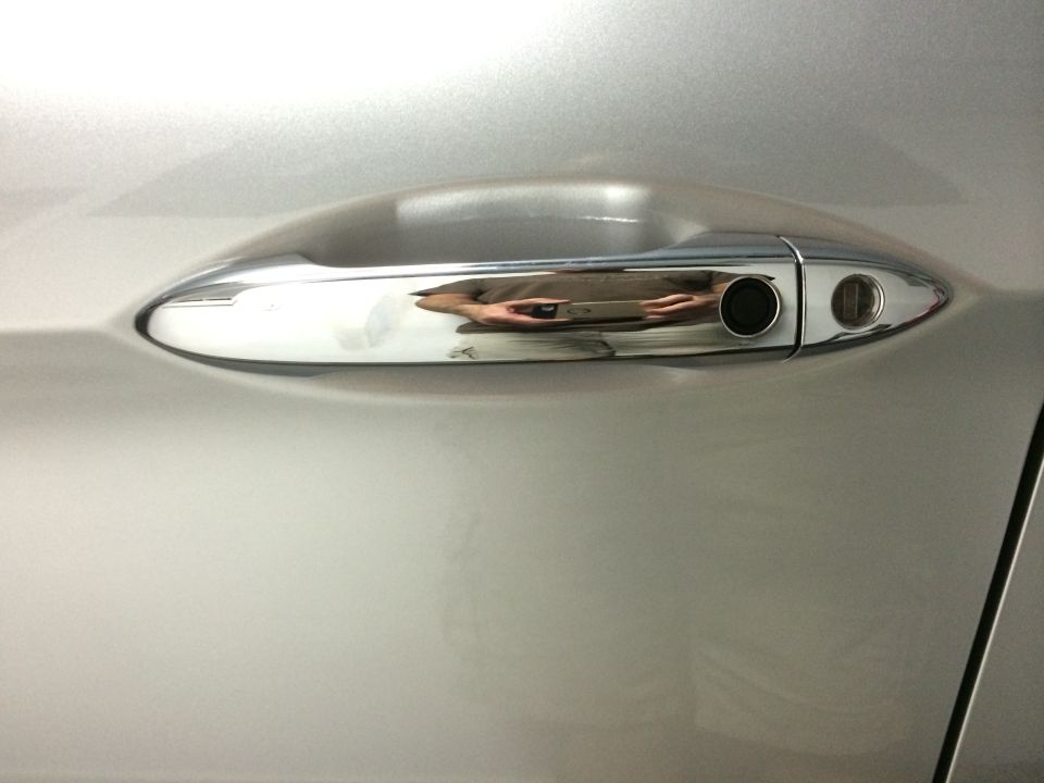 Close-up of a silver car's chrome door handle, showing a keyhole and a reflection of a person holding a phone