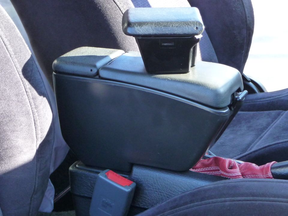 Black plastic armrest with a small storage compartment on top, situated between car seats. Red stitching visible on a fabric strap