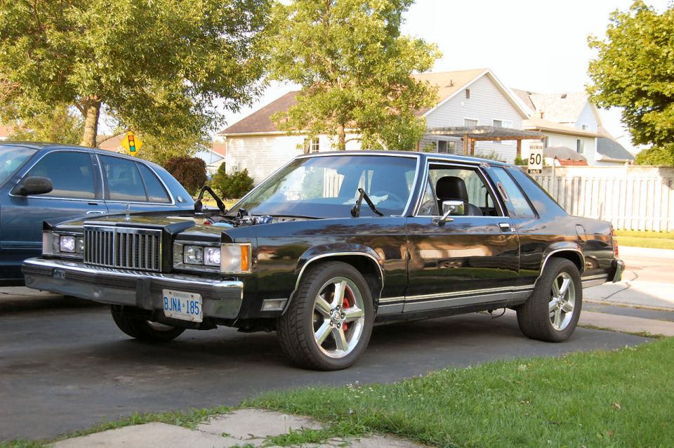 Black coupe with chrome grille and aftermarket wheels parked on driveway. Hood is open