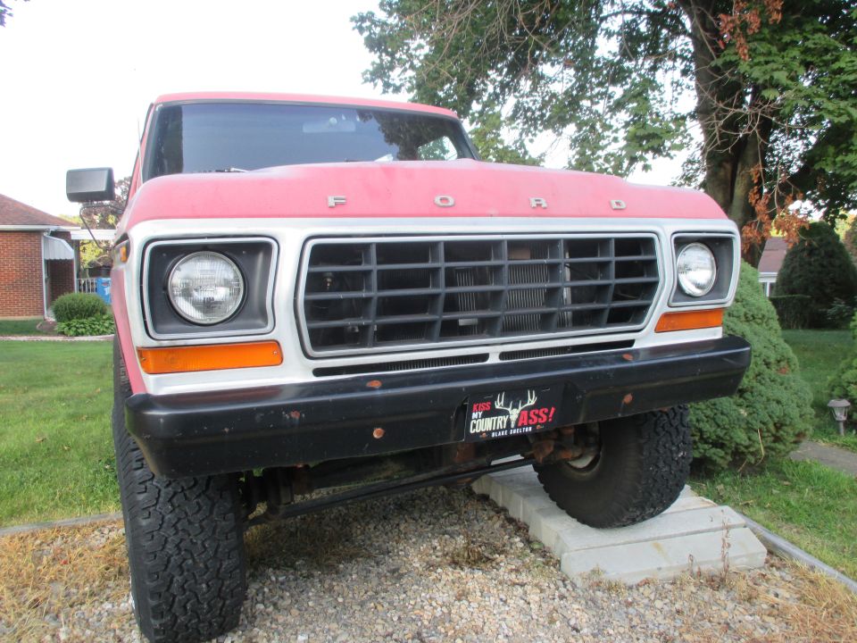 Front view of a red 1979 Ford Bronco with a black grille and a 'Kiss My Country Ass!' bumper sticker