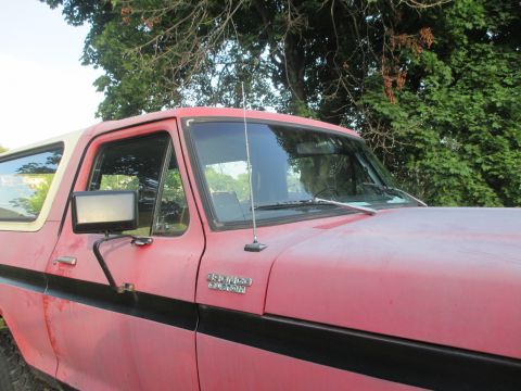 1979 Ford Bronco - A weathered red Ford Bronco with a white top is viewed from the front three-quarter angle.