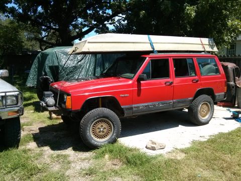 1986 Ford Bronco - A red Jeep Cherokee is parked outdoors with a boat secured to its roof rack.